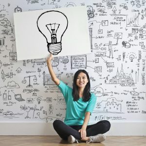 a girl in front of a wall marked with ideas and she is pointing at an large icon of a light bulb