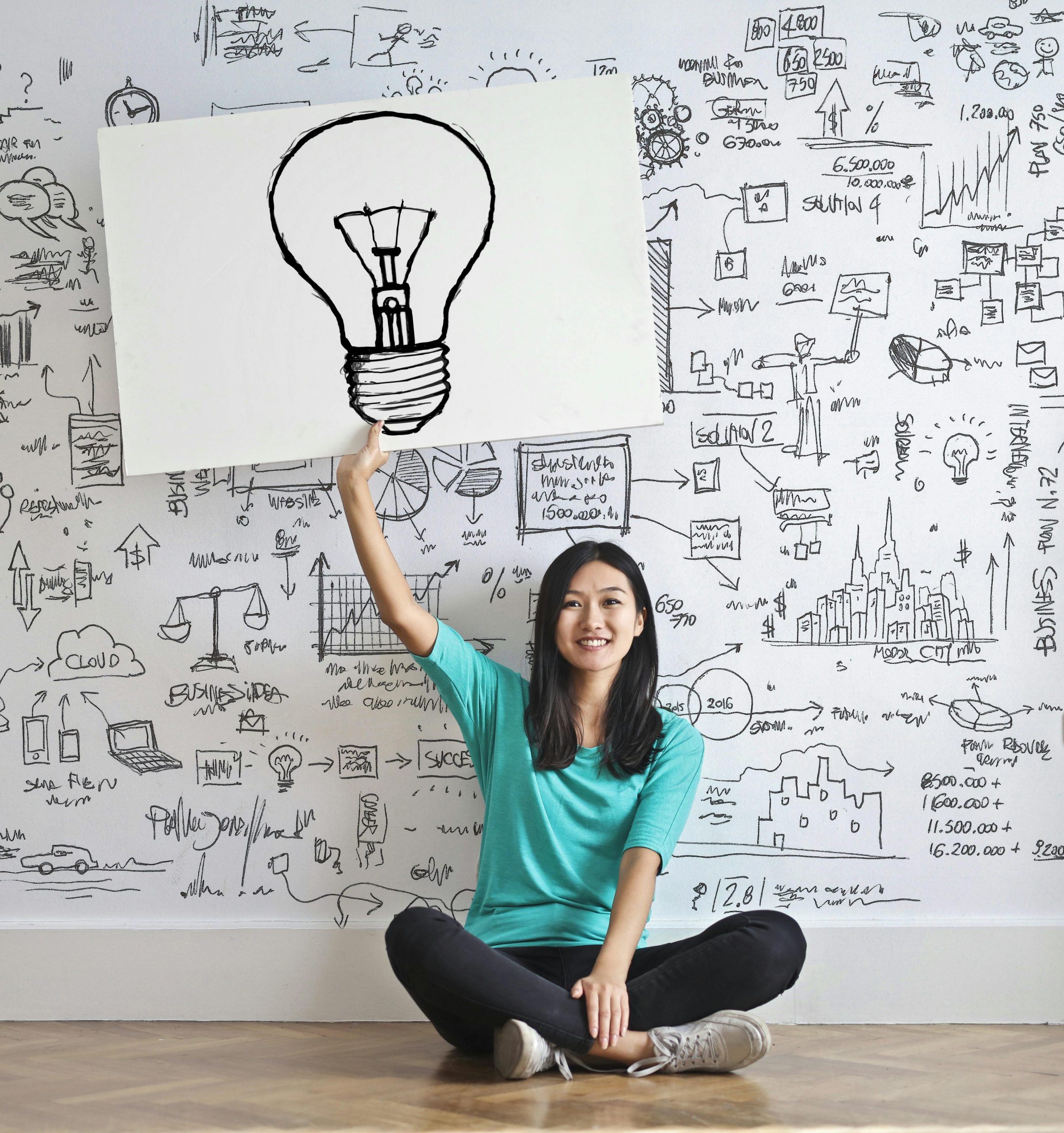 a girl in front of a wall marked with ideas and she is pointing at an large icon of a light bulb